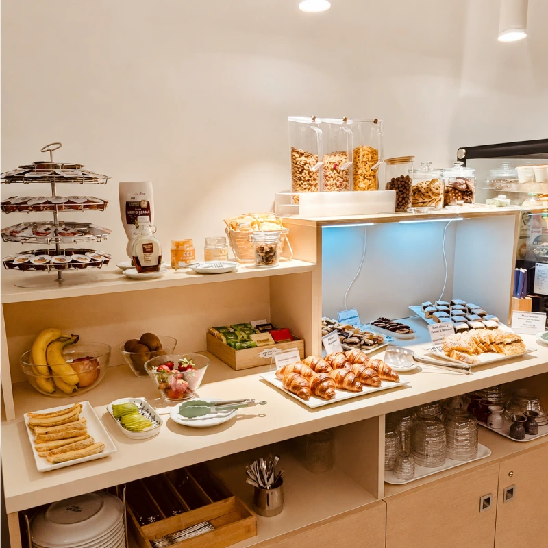Tanti ingredienti da colazione come cornetti, marmellate, frutta, cereali, posate, bicchieri e piatti.
